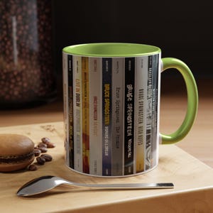 May include: A ceramic mug with a lime green interior and handle, featuring a wrap-around graphic of various Bruce Springsteen album spines. The design includes titles like Live in Dublin, Magic, Working on a Dream, The Promise, and High Hopes.
