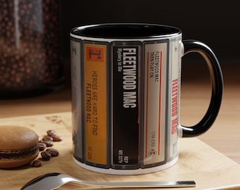 Puede incluir: Una taza de cerámica con interior y asa negros, que presenta un diseño gráfico envolvente de casetes vintage. Las etiquetas muestran texto que incluye Fleetwood Mac, Heroes Are Hard To Find, Mystery to Me y Then Play On.