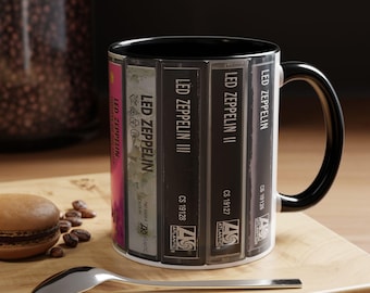 Puede incluir: Una taza de cerámica blanca con interior y asa negros, que presenta un estampado gráfico de varios lomos de casetes de Led Zeppelin. El diseño muestra el texto Led Zeppelin, Led Zeppelin II, Led Zeppelin III y los logotipos de Atlantic.