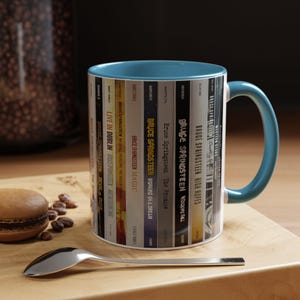 May include: A ceramic mug with a teal interior and handle, featuring a wrap-around graphic of various Bruce Springsteen album spines. The design includes titles like Live in Dublin, Magic, The Promise, Wrecking Ball, High Hopes, and Western Stars.