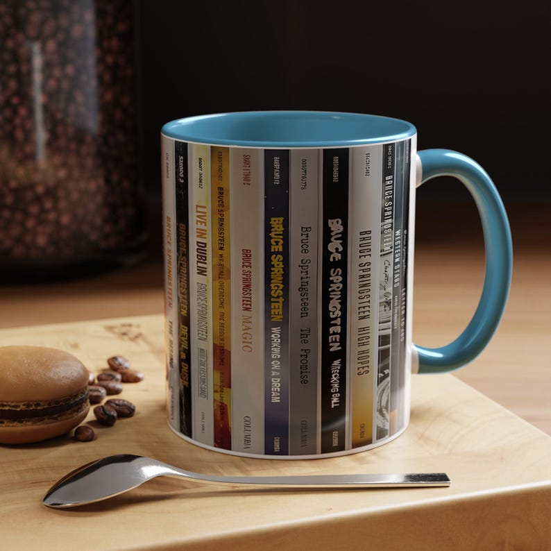 May include: A ceramic mug with a teal interior and handle, featuring a wrap-around graphic of various Bruce Springsteen album spines. The design includes titles like Live in Dublin, Magic, The Promise, Wrecking Ball, High Hopes, and Western Stars.