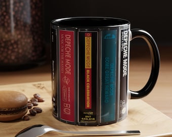 Puede incluir: Una taza de cerámica negra con un estampado gráfico de varias cintas de casete de Depeche Mode dispuestas verticalmente. El diseño incluye títulos de álbumes como Music for the Masses, Black Celebration, Some Great Reward y Speak & Spell.