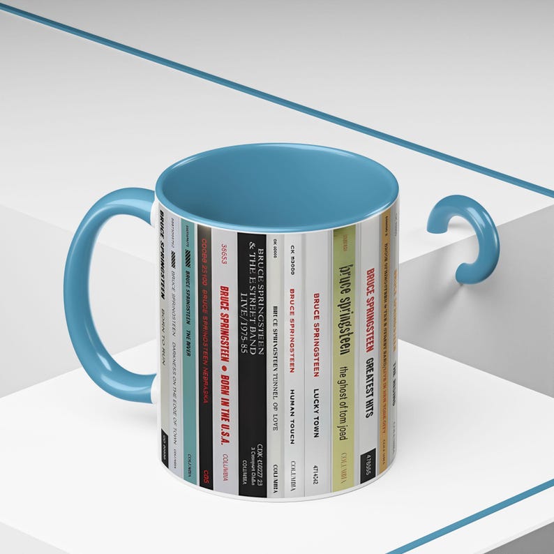 May include: A ceramic mug with a light blue handle and interior. The exterior features a vertical graphic print of various Bruce Springsteen album spines, including titles like Born to Run, The River, Born in the U.S.A., and Greatest Hits.