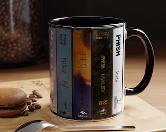 May include: A white ceramic mug with a black interior and handle, featuring a graphic print of multiple cassette tape spines labeled with Phish album titles including Hoist, Rift, Lawn Boy, and Junta, along with the band name Phish printed vertically.