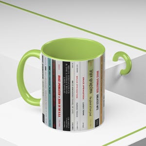 May include: A ceramic mug with a lime green interior and handle. The exterior features a vertical graphic print of various Bruce Springsteen album spines, including titles like Born in the U.S.A., Born to Run, and The River, with visible text and logos.
