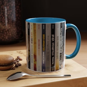 May include: A ceramic mug with a bright blue interior and handle, featuring a wrap-around graphic of various Bruce Springsteen album spines including Live in Dublin, Magic, Working on a Dream, The Promise, Wrecking Ball, High Hopes, and Western Stars.