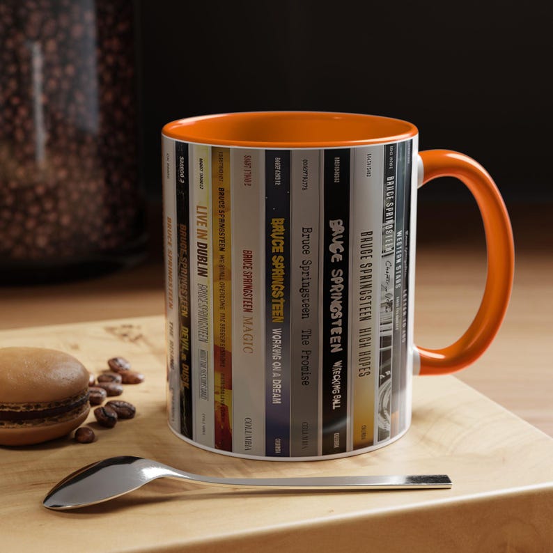 May include: A ceramic mug with a bright orange interior and handle, featuring a wrap-around graphic design of various Bruce Springsteen album spines. The vertical text includes titles like Live in Dublin, Magic, The Promise, and High Hopes.