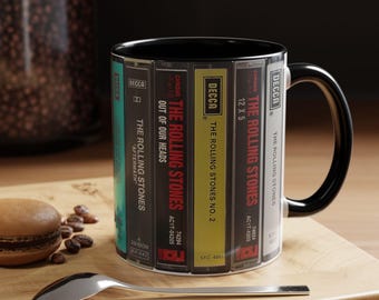 Puede incluir: Una taza de cerámica con interior y asa de color negro, que presenta un diseño envolvente de cintas de casete antiguas etiquetadas con títulos de álbumes de The Rolling Stones como Aftermath, Out of Our Heads y No. 2, junto al logo de Decca.
