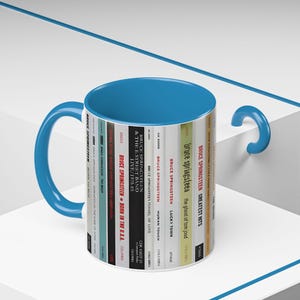 May include: A ceramic mug with a bright blue interior and handle, featuring a wrap-around graphic print of various Bruce Springsteen album spines. The design displays titles like Born to Run, The River, Born in the U.S.A., and Greatest Hits.