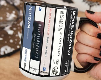 Puede incluir: Una taza de cerámica en blanco y negro con un diseño gráfico de cinco lomos de casetes. El texto incluye Motown, Michael McDonald, Blink of an Eye, Take it to Heart, No Lookin' Back y If That's What It Takes junto a varios logotipos.