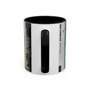 May include: A white ceramic mug with a black interior and handle, featuring a vertical graphic design of Bruce Springsteen album spines. The text includes titles like Born to Run, Western Stars, and Darkness on the Edge of Town in black lettering.