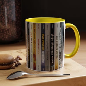 May include: A ceramic mug with a bright yellow interior and handle, featuring a wrap-around graphic of various Bruce Springsteen album spines. The design includes titles like Live in Dublin, Magic, Working on a Dream, The Promise, and High Hopes.