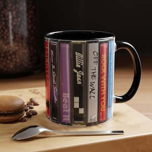 Peut inclure: Un mug en céramique avec une anse et un intérieur noirs, orné d'un motif graphique représentant des cassettes audio verticales. Les étiquettes affichent les textes Thriller, Wanna Be Startin' Somethin', Beat It, Billie Jean, Off The Wall, Rock With You et Don't Stop Till You Get Enough.