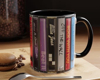 May include: A ceramic mug with a black handle and interior, featuring a wrap-around graphic design of vertical cassette tapes. The labels display text including Thriller, Wanna Be Startin' Somethin', Beat It, Billie Jean, Off The Wall, Rock With You, and Don't Stop Till You Get Enough.