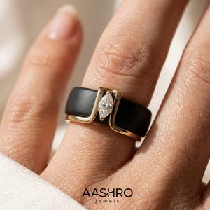 May include: A matte black band ring featuring a gold-toned metal inlay that forms a vertical setting for a single marquise-cut clear gemstone. The ring is positioned on a finger, and the text AASHRO jewels is visible at the bottom of the image.