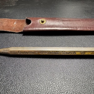 May include: A metallic mechanical pencil with a textured hexagonal barrel and a pointed tip. The barrel features the engraved text Gummi-Wollstatt, Zürich and a small circular logo. A brown leather protective sleeve with a brass snap button is included.