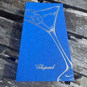 May include: A rectangular blue box featuring a gold-toned line art illustration of a martini glass containing two dice. The brand name Chopard is printed in gold script lettering near the bottom center of the box surface.