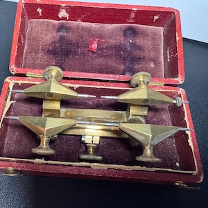 May include: A brass watchmaker's staking tool set featuring four pointed metal stakes with knurled adjustment knobs, housed in a rectangular red case with a dark purple velvet interior lining and worn edges on the box frame.