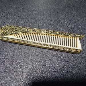 May include: A folding hair comb featuring an off-white plastic comb insert housed within a decorative gold-tone metal case. The metal exterior is engraved with a detailed floral pattern and leaf motifs along the entire length of the handle.