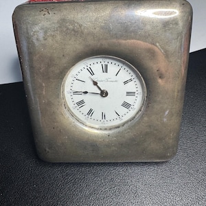 May include: A square-shaped silver-toned metal desk clock with a rounded indentation in the center. The white clock face features black Roman numerals, thin black hands, and the text Federico Fornotti printed in black script across the center.