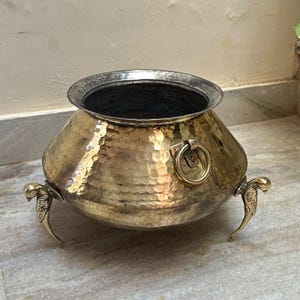 Pode incluir: Um pote redondo de latão martelado com uma abertura interior larga e escura. O exterior apresenta uma superfície texturizada com covinhas, duas alças laterais em forma de anel e três pernas decorativas em forma de pássaro.