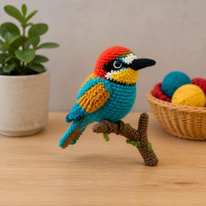 Op de afbeelding: Een gehaakt bijeneter-vogelbeeldje met een levendige rode kroon, een zwart oogmasker, een felgele keel en een blauw lichaam met oranje vleugelaccenten. De vogel zit op een bruine gehaakte tak met kleine groene bladdetails.