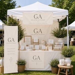Puede incluir: Un puesto exterior con toldo blanco y carteles color crema con el texto negro GA GRAPHICSSHOP. La exhibición incluye artículos de papelería, pequeñas cajas de regalo y plantas verdes en cestas de mimbre y jarras blancas.