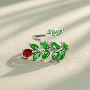 Könnte beinhalten: Ein silberfarbener offener Ring mit einem Zweigdesign und mehreren marquise-geschliffenen grünen Steinen, die wie Blätter aussehen. Ein Ende des Rings zeigt einen birnenförmigen roten Stein, während das andere Ende ein kleines Blatt mit grünen Pavé-Steinen aufweist.