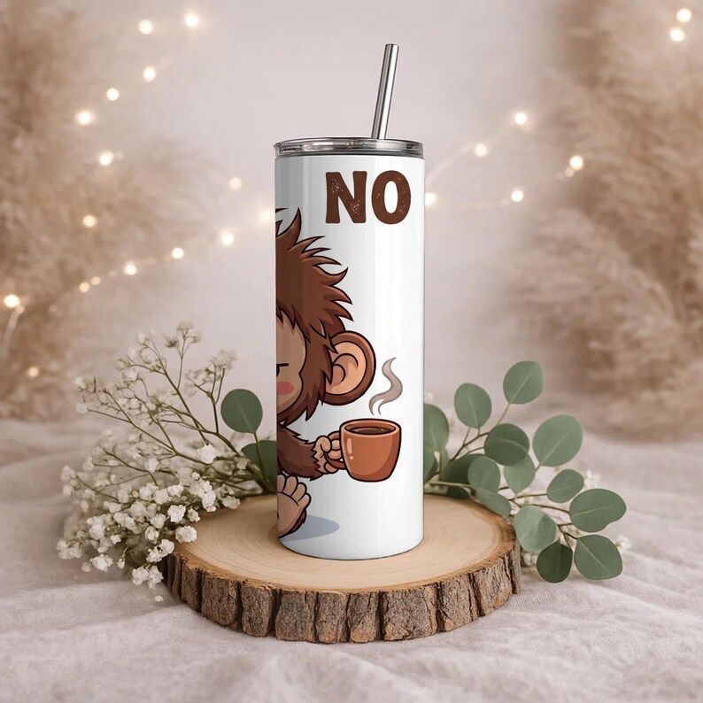Op de afbeelding: Een witte cilindrische beker met een metalen rietje met een cartoonillustratie van een bruine aap die een dampende bruine koffiemok vasthoudt. Het woord NO is in grote bruine hoofdletters boven de aap gedrukt. De beker heeft een deksel.