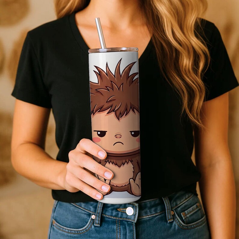 Op de afbeelding: Een hoge cilindrische beker met een metalen rietje, voorzien van een grafische illustratie van een bruin, harig cartoonachtig wezen met warrig haar, een norse gezichtsuitdrukking en roze wangen tegen een lichtgrijze achtergrond.