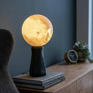 Può includere: Una lampada sferica a forma di luna con una superficie ruvida e luminosa che emette una calda luce gialla. La lampada è sostenuta da una solida base cilindrica affusolata di colore nero opaco, appoggiata su due libri intitolati The Moon: An Artistic Journey.