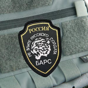 May include: A black shield-shaped patch with a gold border, featuring a white tiger head graphic in the center. The text reads "РОССИЯ", "НЕ ЗЛИТЕ РУССКОГО СОЛДАТА", and "БАРС" in white and gold Cyrillic lettering, attached to a grey tactical vest.