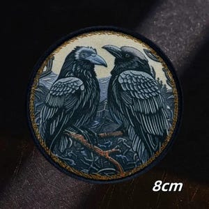 Könnte beinhalten: Ein kreisförmiger bestickter Aufnäher mit zwei schwarzen Raben, die auf einem Ast vor einer gedämpften Landschaft sitzen. Das Motiv ist von einem goldfarbenen Rand umgeben und enthält den Text 8cm, der einen Durchmesser von 8 cm angibt.