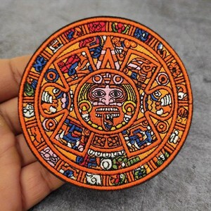 Puede incluir: Un parche bordado circular con un diseño detallado de la piedra del sol azteca. El bordado utiliza hilos vibrantes de color naranja, amarillo, azul y blanco para representar un rostro central rodeado de complejos patrones geométricos.