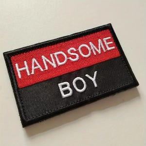 May include: A rectangular embroidered patch featuring a split design with a red top section and a black bottom section. The word HANDSOME is stitched in white thread on the red background, and the word BOY is stitched in white on the black.