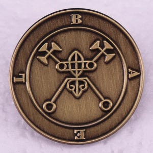 Könnte beinhalten: Eine kreisförmige Anstecknadel aus bronzefarbenem Metall mit einem geprägten Siegel in der Mitte. Das Design zeigt ein zentrales geometrisches Emblem mit gekreuzten Linien und kreisförmigen Enden, umgeben von den Buchstaben B, L, A und E.