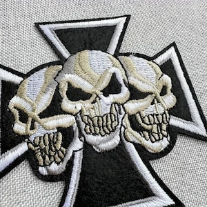 May include: An embroidered patch featuring three skulls arranged horizontally, rendered in shades of white, gray, and tan thread. The skulls are positioned over a black iron cross emblem with a thick white border, creating a high-contrast design.