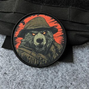 以下が含まれることがあります： つばの広い帽子をかぶったハイイログマが描かれた円形の刺繍ワッペン。クマは光るオレンジ色の目をし、口を開けており、鮮やかなオレンジと黒のサンバーストの背景に、太い黒のステッチの縁取りが施されています。