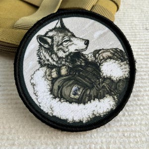 May include: A circular embroidered patch featuring a detailed illustration of a wolf wearing tactical gear. The wolf is depicted in shades of gray and black, set against a white background with a thick black border. The design shows the wolf looking to the right, wearing a vest with visible straps and a small rectangular patch on the chest area.