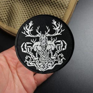 May include: A circular black embroidered patch featuring a white thread design of a stylized deer skull with large, intricate antlers and skeletal elements. The embroidery is dense and detailed, showcasing a symmetrical composition on a dark fabric base.