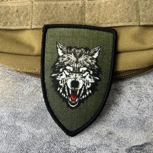 Puede incluir: Un parche en forma de escudo con una ilustración detallada de una cabeza de lobo gruñendo con pelaje blanco, gris y negro. El lobo tiene la boca abierta mostrando encías rojas y dientes afilados sobre un fondo de tela verde oliva.
