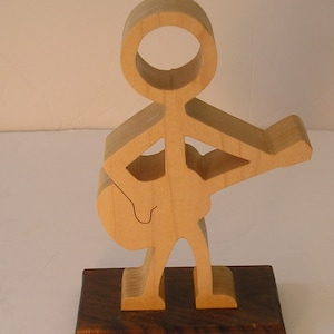 May include: Wooden figurine of a person playing a guitar. The figurine is made of light wood and is standing on a dark wood base.