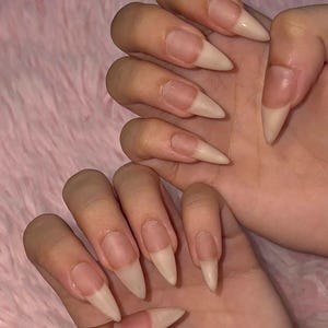 May include: Two hands displaying long, almond-shaped artificial nails with a natural, nude-toned finish. The nails feature a clear base near the cuticle that transitions into a solid, opaque beige color toward the pointed tips, creating a clean look.