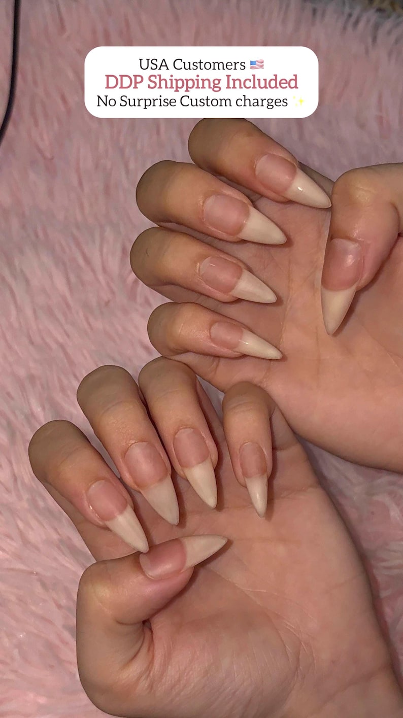 May include: Two hands displaying long, almond-shaped artificial nails with a natural, nude-toned finish. The nails feature a clear base near the cuticle that transitions into a solid, opaque beige color toward the pointed tips, creating a clean look.
