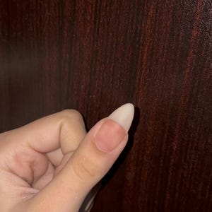 May include: A single almond-shaped fingernail with a natural, unpainted appearance. The nail plate is smooth and light-toned, extending past the fingertip with a clean, rounded edge. The surrounding skin is light-toned and appears healthy.