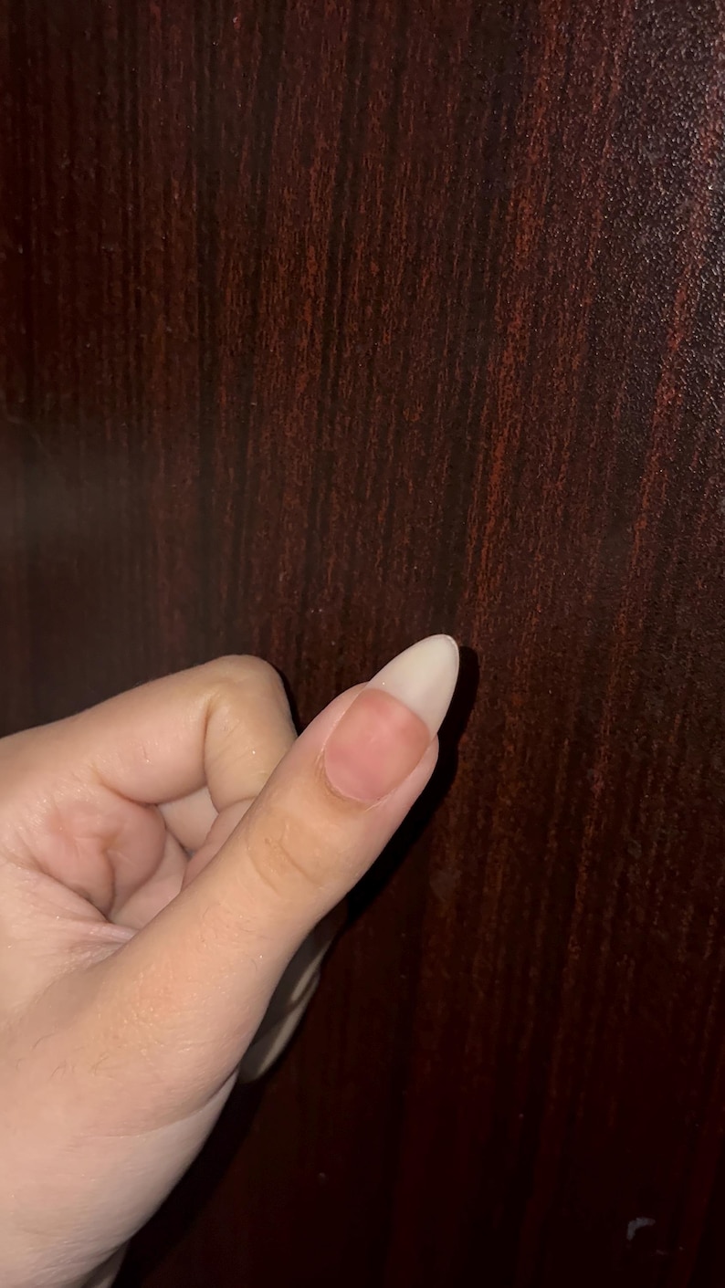 May include: A single almond-shaped fingernail with a natural, unpainted appearance. The nail plate is smooth and light-toned, extending past the fingertip with a clean, rounded edge. The surrounding skin is light-toned and appears healthy.