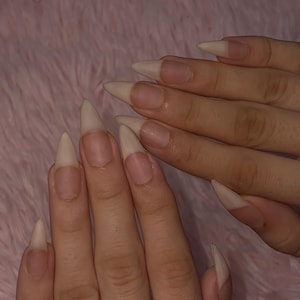 May include: Long, pointed stiletto-shaped fingernails with a natural, translucent beige finish. The nails feature a clean, unpainted appearance with a smooth, tapered edge that extends from the nail bed, showcasing a sharp, elongated silhouette.