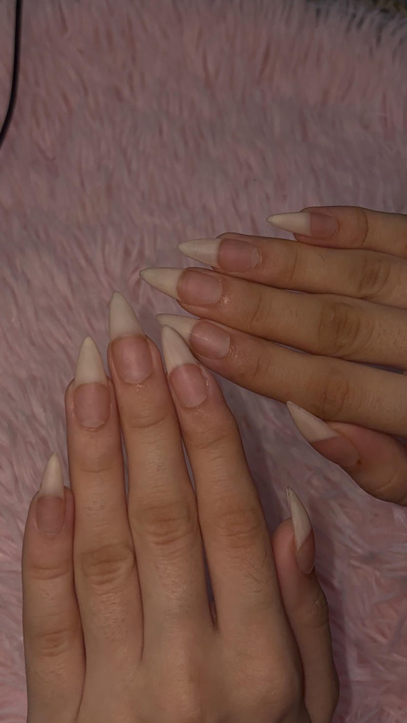 May include: Long, pointed stiletto-shaped fingernails with a natural, translucent beige finish. The nails feature a clean, unpainted appearance with a smooth, tapered edge that extends from the nail bed, showcasing a sharp, elongated silhouette.