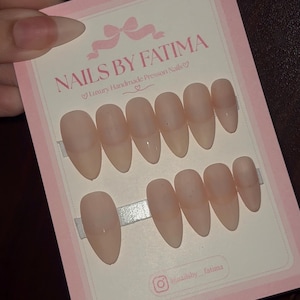 May include: A set of ten almond-shaped, nude-colored press-on nails displayed on a rectangular pink and white card. The card features the text NAILS BY FATIMA, Luxury Handmade Presson Nails, and an Instagram handle, all printed in a dark pink font.