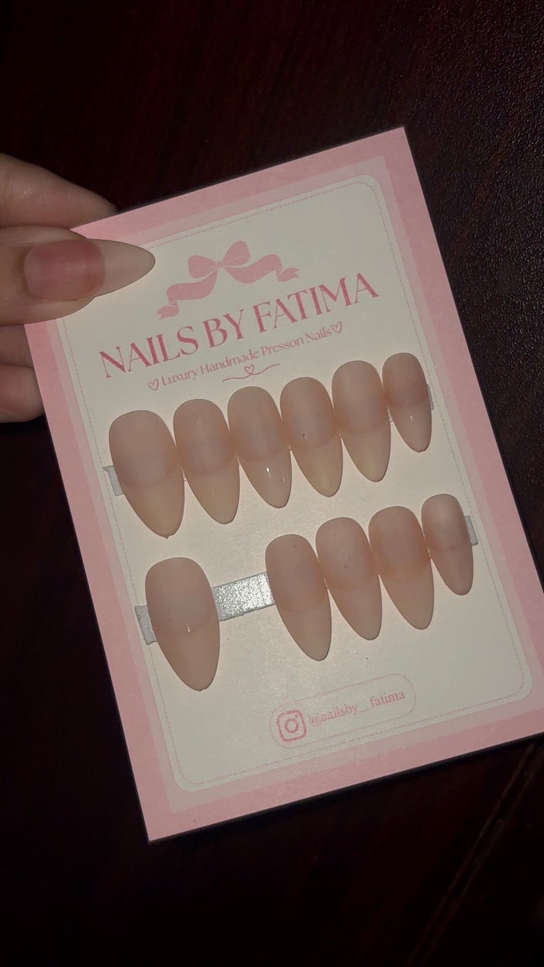 May include: A set of ten almond-shaped, nude-colored press-on nails displayed on a rectangular pink and white card. The card features the text NAILS BY FATIMA, Luxury Handmade Presson Nails, and an Instagram handle, all printed in a dark pink font.